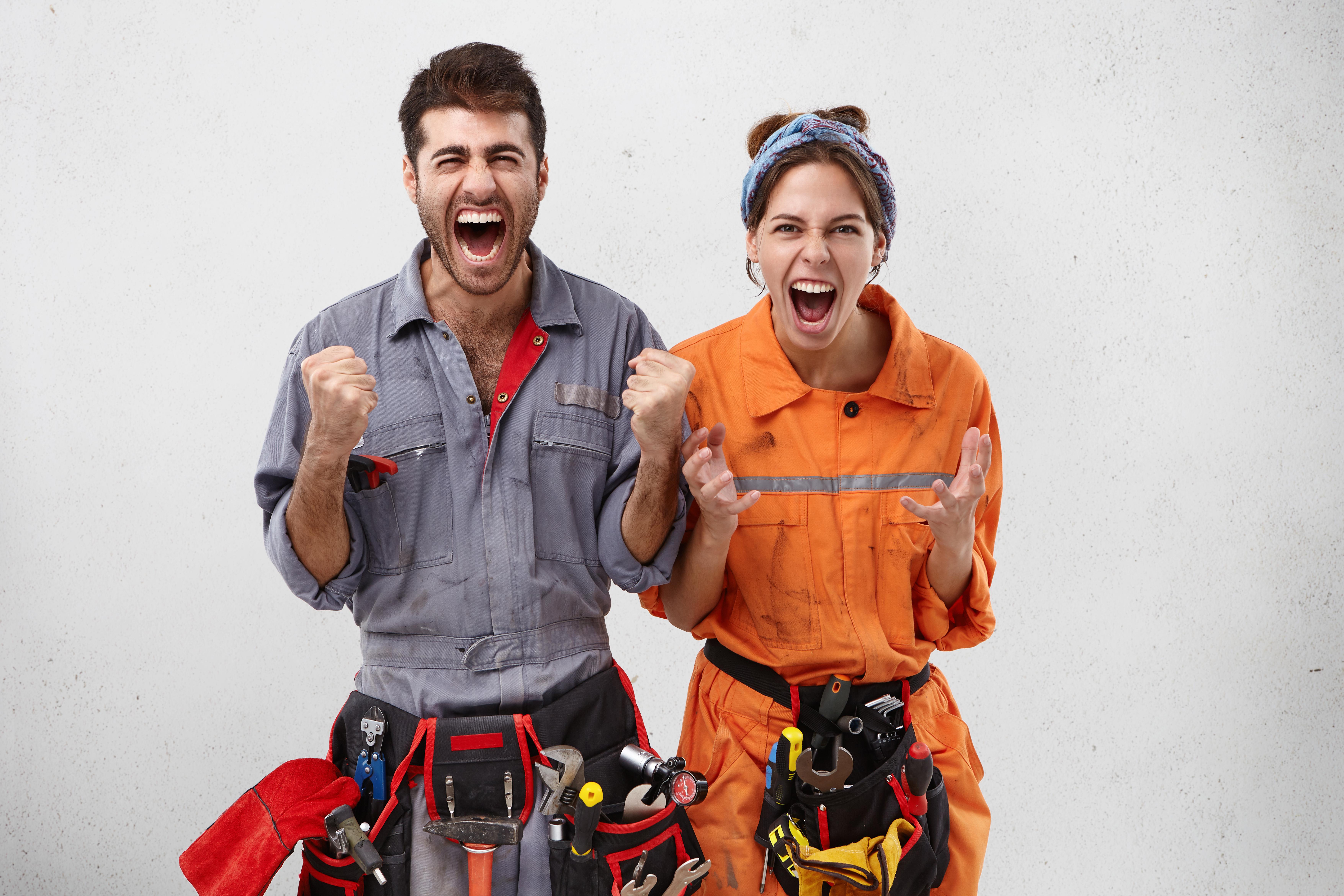 Overjoyed successful female and male maintenance workers clench fists, shout with excitement, being glad to finish work in advance. Emotional bearded handyman and his colleague gestures indoors